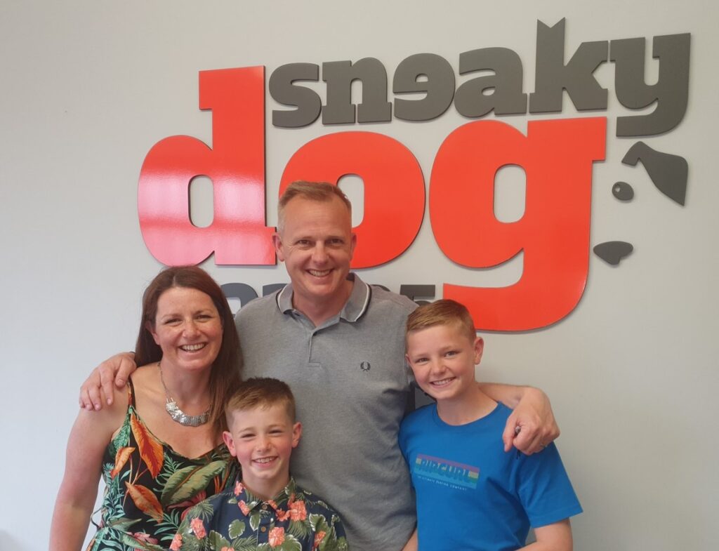 A happy family posing in front of the Sneaky Dog Escapes logo enjoying a Father's Day experience. There is a father, mother and 2 sons. 