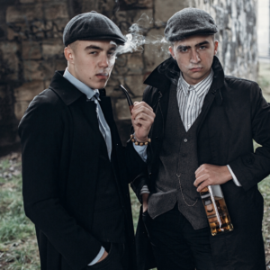 2 men dress in 1930s clothing. Smoking and drinking whisky. Dressed up for an escape room as a stag do activity.